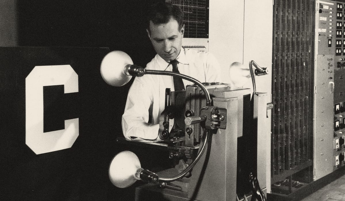 A man at the Mark I Perceptron, holding a large letter C up to the machine's lamps; racks of patch wiring behind him.